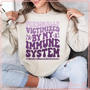 Puede incluir: Sudadera color crema con el texto morado "PERSONALLY VICTIMIZED BY MY IMMUNE SYSTEM". El diseño incluye lazos morados y el hashtag "#Crohn'sDiseaseAwareness". La sudadera es de estilo casual.