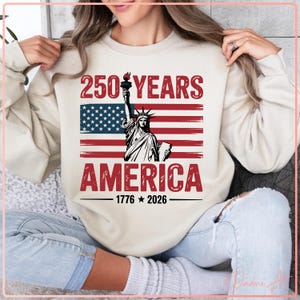 May include: Cream-colored sweatshirt featuring a graphic of the Statue of Liberty holding a torch, set against a red and blue American flag design. The text reads "250 YEARS AMERICA 1776-2026".