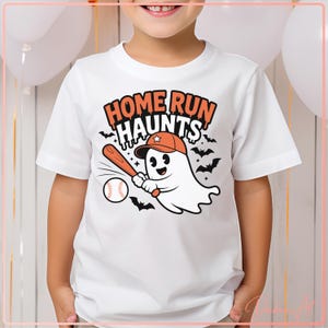 May include: White t-shirt with a cartoon ghost wearing an orange baseball cap and holding a bat, ready to hit a baseball. The shirt has the text "HOME RUN HAUNTS" in orange and black, with bats in the background.