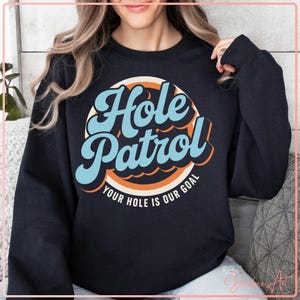 以下が含まれることがあります： レトロなフォントで「Hole Patrol」の文字が入った黒いスウェットシャツ。スローガン「Your Hole Is Our Goal」が円形デザインで配置されています。テキストはライトブルー、オレンジ、白です。