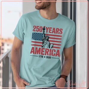 May include: Light blue t-shirt featuring a graphic of the American flag with the Statue of Liberty. The text reads "250 YEARS AMERICA 1776-2026". The design is in red, white, and blue.