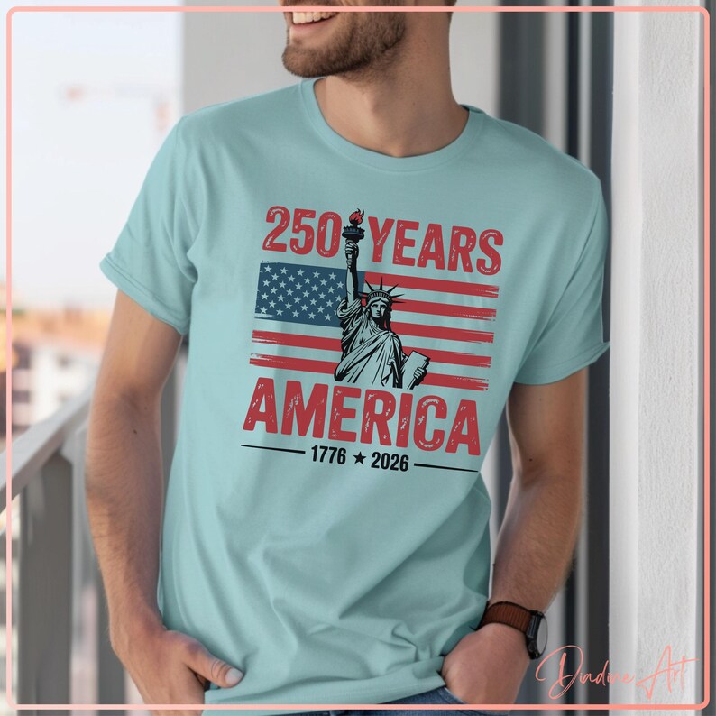 May include: Light blue t-shirt featuring a graphic of the American flag with the Statue of Liberty. The text reads "250 YEARS AMERICA 1776-2026". The design is in red, white, and blue.