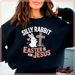 Puede incluir: Sudadera negra con el texto "SILLY RABBIT EASTER IS FOR JESUS". El diseño incluye un conejito blanco, una cruz y dos corazones rosas. El texto y los gráficos son en blanco y rosa.