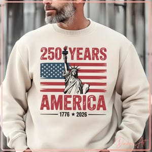 May include: Cream-colored sweatshirt featuring a graphic of the Statue of Liberty, an American flag, and the text "250 YEARS AMERICA 1776-2026". The design is in red, white, and blue.