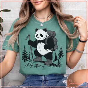 May include: Green t-shirt with a black and white panda bear design. The panda wears a backpack and holds a hiking stick, set against a backdrop of pine trees. The shirt has a relaxed fit.