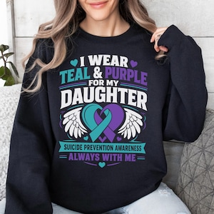 Puede incluir: Una sudadera negra de cuello redondo con el texto I WEAR TEAL & PURPLE FOR MY DAUGHTER. El diseño incluye un lazo de concienciación turquesa y morado en forma de corazón con alas de ángel blancas y el texto SUICIDE PREVENTION AWARENESS ALWAYS WITH ME.