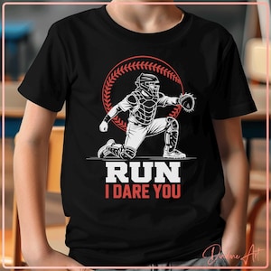 May include: Black t-shirt featuring a baseball catcher graphic in white and red, kneeling in a defensive stance. The text "RUN I DARE YOU" is displayed below the graphic, set against a baseball-themed background.