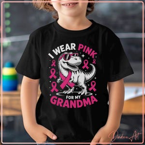 Puede incluir: Camiseta negra con un dinosaurio rosa con gafas de sol y una cinta rosa. El texto dice "I WEAR PINK FOR MY GRANDMA" en rosa y blanco. La camiseta la lleva un niño.