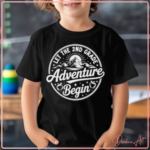 May include: Black t-shirt featuring a white circular design with the text "Let the 2nd Grade Adventure Begin." The graphic includes a mountain, apples, and stars. The shirt is worn by a child.