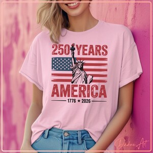 May include: A light pink t-shirt with the text "250 YEARS AMERICA 1776-2026" and an illustration of the Statue of Liberty in front of an American flag. The shirt is a casual style.