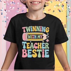 May include: Black t-shirt with the text "Twinning with my Teacher Bestie" in colorful, retro-style lettering. A large pencil graphic is featured in the center of the design. The shirt is worn by a child.
