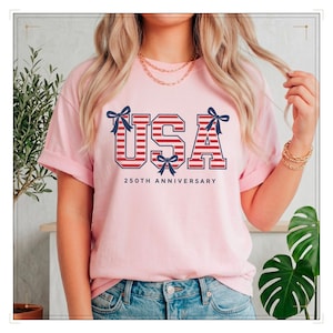 Könnte beinhalten: Ein hellrosa T-Shirt mit dem Wort "USA" in rot-weiß gestreiften Buchstaben, akzentuiert mit marineblauen Schleifen. Der Text "250TH ANNIVERSARY" steht unter dem Wort "USA".