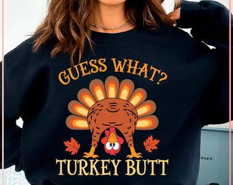 Może przedstawiać: Czarna bluza z napisem "GUESS WHAT? TURKEY BUTT" w kolorze pomarańczowym i żółtym. Grafika przedstawia kreskówkowego indyka z tyłem do góry, z jesiennymi liśćmi po obu stronach.