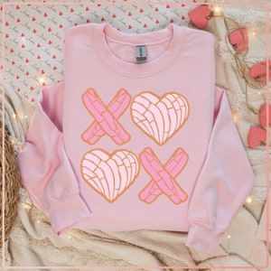 May include: A pink sweatshirt with a graphic design of two pink hearts and two pink X's. The hearts are in the shape of a traditional Mexican sweet bread called a concha.