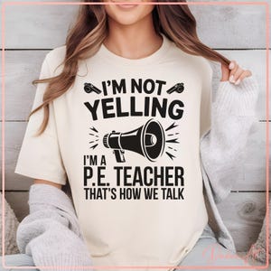 May include: White t-shirt with black text that reads "I'm not yelling I'm a P.E. teacher that's how we talk". The text is accompanied by a black and white illustration of a megaphone.