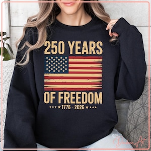 Pode incluir: Um moletom azul marinho com o texto "250 YEARS OF FREEDOM" e um design da bandeira americana. O design inclui os anos 1776-2026 e é impresso em estilo vintage. O moletom é uma peça de roupa casual.