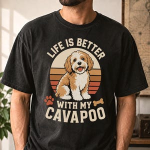 Könnte beinhalten: Ein schwarzes T-Shirt mit kurzen Ärmeln und einer Grafik eines braun-weißen Cavapoo-Hundes, der vor einem Sonnenuntergangskreis sitzt. Der Text lautet "LIFE IS BETTER WITH MY CAVAPOO", ergänzt durch eine Pfotenabdruck- und eine Knochengrafik.
