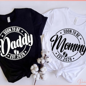 May include: Black and white t-shirts with the text "SOON TO BE Daddy EST. 2026" and "SOON TO BE Mommy EST. 2026" in a circular design. The shirts are a pregnancy announcement theme.