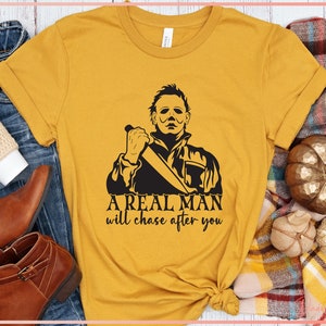 May include: A mustard yellow t-shirt with a black graphic of a man holding a knife. The text "A real man will chase after you" is printed below the graphic.