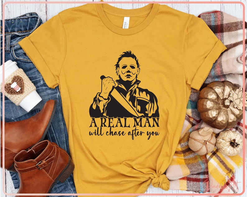 May include: A mustard yellow t-shirt with a black graphic of a man holding a knife. The text "A real man will chase after you" is printed below the graphic.