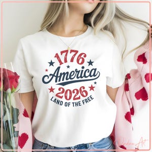 Op de afbeelding: Wit T-shirt met een patriottisch ontwerp. Het ontwerp toont de tekst "1776 America 2026 Land of the Free" in rood en marineblauw, omringd door sterren. Het shirt is een klassieke ronde hals.