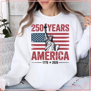 May include: White sweatshirt featuring a graphic design celebrating 250 years of America. The design includes the Statue of Liberty, the American flag, and the text "250 YEARS AMERICA 1776-2026" in red and black.