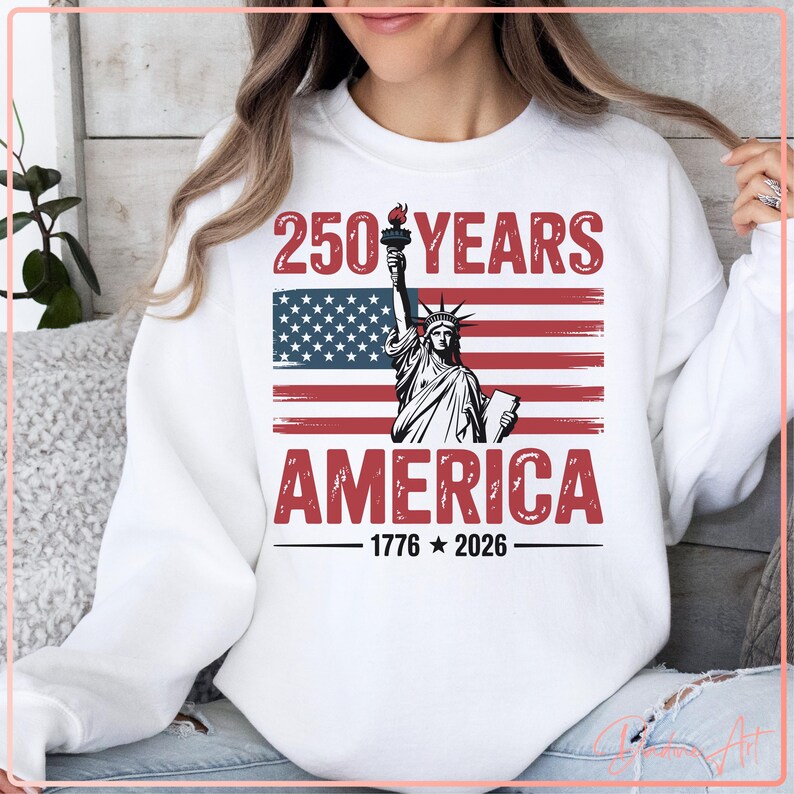 May include: White sweatshirt featuring a graphic design celebrating 250 years of America. The design includes the Statue of Liberty, the American flag, and the text "250 YEARS AMERICA 1776-2026" in red and black.