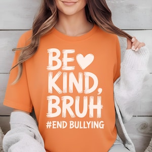 May include: Orange t-shirt with the words "BE KIND, BRUH" in white, brush-style lettering. A white heart replaces the "O" in "BE". The phrase "#END BULLYING" is printed below. The shirt is a casual wear item.