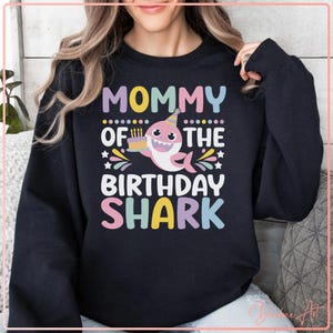 Puede incluir: Sudadera negra con el texto "MOMMY OF THE BIRTHDAY SHARK" en letras coloridas. Un tiburón de dibujos animados con un gorro de fiesta y sosteniendo un pastel está en el centro del diseño.