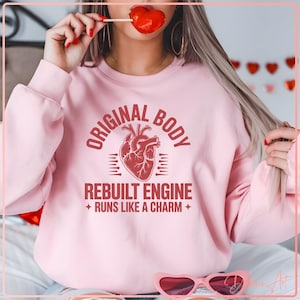 May include: Light pink sweatshirt with the text "ORIGINAL BODY REBUILT ENGINE RUNS LIKE A CHARM" and a red anatomical heart graphic. The sweatshirt is a crew neck style.
