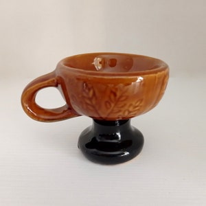 May include: A small brown ceramic cup with a handle and a black base. The cup has a leaf design.