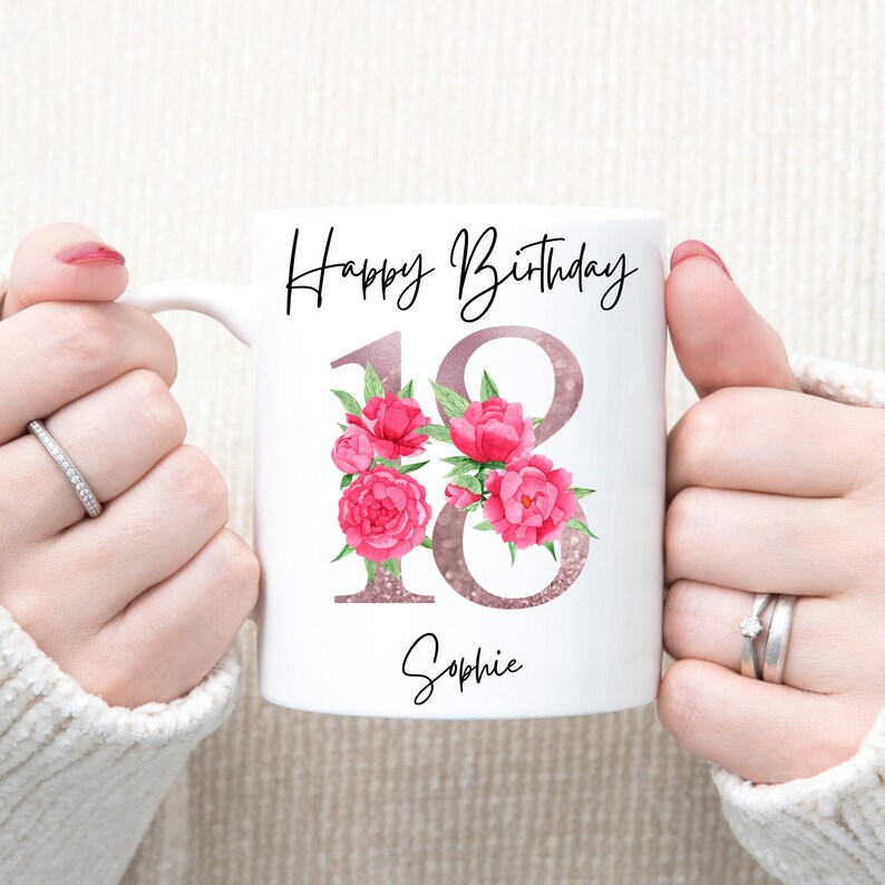 Puede incluir: Taza de cer&aacute;mica blanca con la frase "Happy Birthday" en escritura negra. El n&uacute;mero "18" tiene un dise&ntilde;o de purpurina oro rosa, adornado con acentos florales rosas. El nombre "Sophie" est&aacute; escrito debajo.