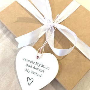 May include: A white ceramic heart-shaped ornament with the text "Forever My Mum And Always My Friend" and a small heart at the bottom. The ornament is tied with a white ribbon and is on a brown gift box.