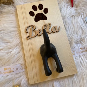 May include: A wooden sign with a black paw print cutout and the name "Bella" written in cursive. A black dog-shaped hook is attached to the sign.