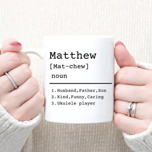 May include: White ceramic mug with a black design that says "Matthew [Mat-chew] noun 1. Husband, Father, Son 2. Kind, Funny, Caring 3. Ukulele player"