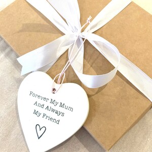May include: A white ceramic heart-shaped ornament with the text "Forever My Mum And Always My Friend" and a small heart design. The ornament is tied with a white ribbon and pink string.