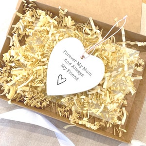 May include: White ceramic heart-shaped ornament with the text "Forever My Mum And Always My Friend" and a small heart symbol. The ornament is in a box with shredded paper.