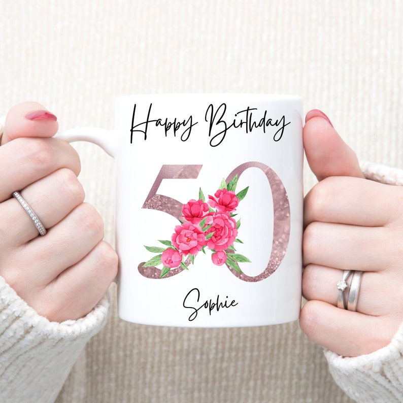 May include: White ceramic mug with a pink glitter design and pink flowers. The mug says "Happy Birthday 50 Sophie".