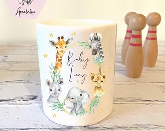 May include: A white ceramic mug with a pink ombre design featuring a giraffe, zebra, rhino, elephant, and lion. The mug has a gold rim and green leaves around the animals. The text "Baby Lucy" is written in cursive on the mug.