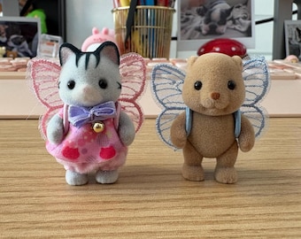 Op de afbeelding: Twee kleine pluche speelgoedjes met vlinder vleugels. Het eerste speelgoed is een grijze kat met roze en witte kleding en een paarse strik. Het tweede speelgoed is een bruine beer met blauwe vleugels.