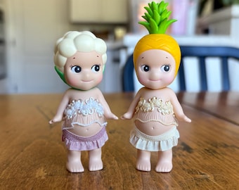 May include: Two small figurines with large eyes and smiling faces. One has a cauliflower-shaped head, the other a pineapple head. Both wear light-colored tops with floral designs and ruffled skirts. The figurines are on a wooden surface.