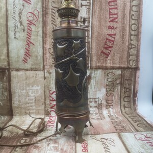 Peut inclure: Une lampe à huile vintage avec une finition en cuivre et un motif décoratif de feuilles et de vignes. La lampe a une base ronde avec trois pieds et un abat-jour cylindrique.