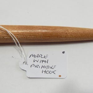 May include: A light brown wooden crochet hook with a rounded knob handle and a metal hook tip. A white rectangular tag is attached with a string, featuring the handwritten text: MAPLE WITH PRIMITIVE HOOK. The tool has a smooth, polished finish.