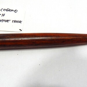 May include: A crochet hook with a dark brown, polished wooden handle featuring a rounded knob at the end. A white tag is attached to the handle with string, displaying the handwritten text: COCOBOLO WITH PRIMITIVE HOOK. The metal hook is silver.