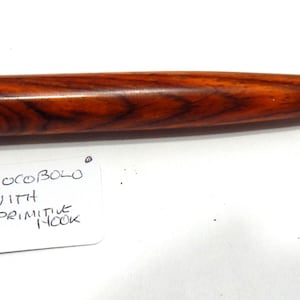 May include: A crochet hook with a polished reddish-brown wood handle featuring a rounded end and a metal hook tip. A white paper tag is attached to the handle with a string, displaying the handwritten text: COCOBOLO WITH PRIMITIVE HOOK.