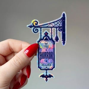 May include: A holographic sticker shaped like a hanging sign with a dark blue outline. The sign features a skull icon, a spiderweb design, and the text "THE HORROR READER" in a stylized font. It is suspended by chains and includes bat-shaped accents.