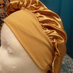 Könnte beinhalten: Eine goldene Satin-Schlafhaube mit gerafftem Oberteil. Die Haube befindet sich auf einem weißen Mannequin-Kopf.