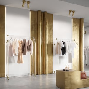 Pode incluir: O interior de uma loja de roupas com paredes douradas e um teto branco. Duas araras de roupas com roupas em cabides são visíveis. Um expositor dourado com um par de sapatos e uma bolsa está em primeiro plano.