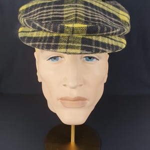 May include: A yellow and black checked newsboy cap. The cap is displayed on a mannequin head with blue eyes and a neutral skin tone. The hat has a classic, rounded shape and a small brim. The background is a dark blue.