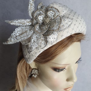 May include: A white felt beret with a large, silver sequined flower embellishment. The flower has a pearl-like center and is accented with silver beads.